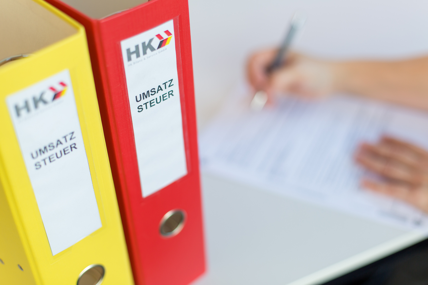 Eine Frau füllt ein Formular aus. Daneben stehen ein gelber und ein roter Ordner mit der Beschriftung Umsatzsteuer.